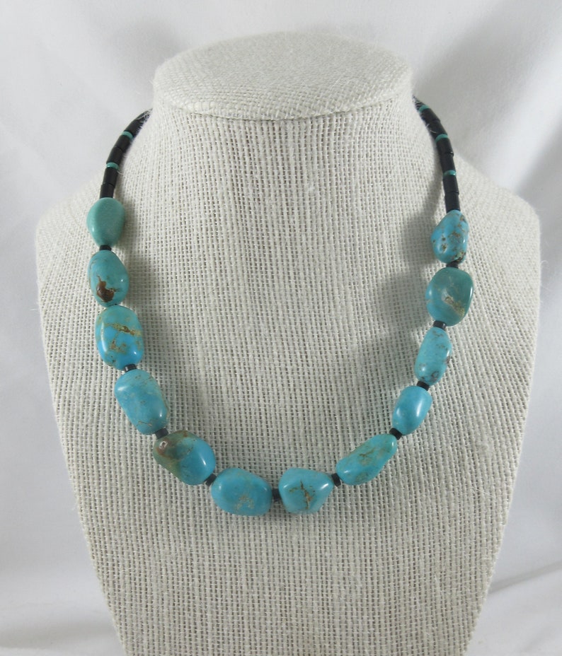 May include: A turquoise and black necklace with large, oval-shaped turquoise beads. The necklace is strung on a black cord.