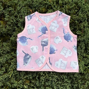 May include: Pink sleeveless vest with a pattern of grey cats carrying shopping bags. The vest has a button closure.