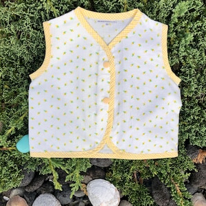 May include: A white vest with a yellow floral pattern and yellow gingham trim. The vest has three yellow buttons and is labeled "HAWAIIANPATCHES" and "XS". The vest is likely for a child.