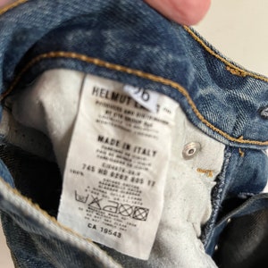 Puede incluir: Una etiqueta blanca con texto negro unida a un par de pantalones vaqueros azules. La etiqueta dice "HELMUT LANG", "MADE IN ITALY", "745 ND #202 605 17", "100%" y "CA 19543".