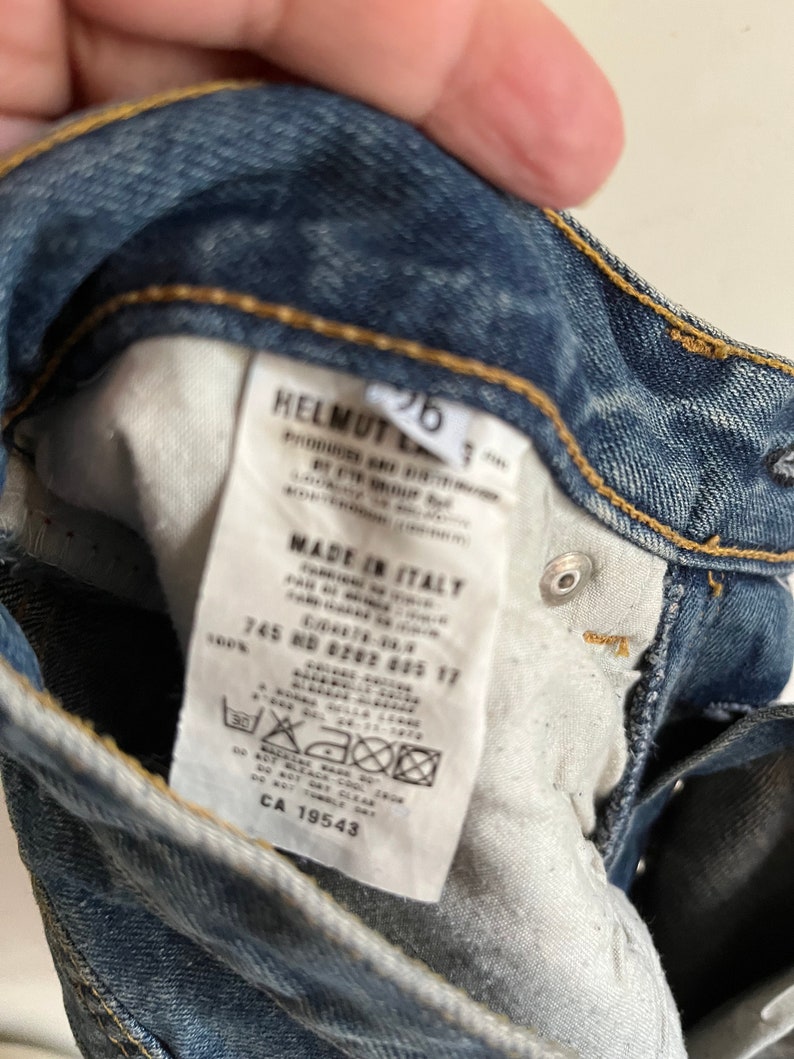 Puede incluir: Una etiqueta blanca con texto negro unida a un par de pantalones vaqueros azules. La etiqueta dice "HELMUT LANG", "MADE IN ITALY", "745 ND #202 605 17", "100%" y "CA 19543".