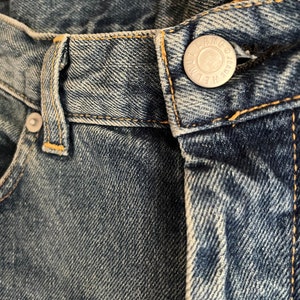 Puede incluir: Primer plano de la cintura de un par de pantalones vaqueros azules con un bot&oacute;n plateado que dice "Wrangler Jeans"