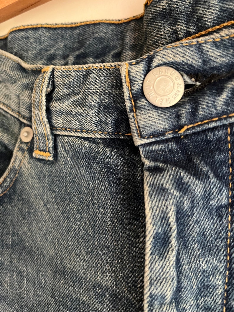 Puede incluir: Primer plano de la cintura de un par de pantalones vaqueros azules con un bot&oacute;n plateado que dice "Wrangler Jeans"