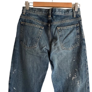 Puede incluir: Un par de pantalones vaqueros azules de mezclilla con salpicaduras de pintura blanca en las piernas. Los vaqueros tienen un estilo cl&aacute;sico de cinco bolsillos y est&aacute;n colgados de una percha de madera.