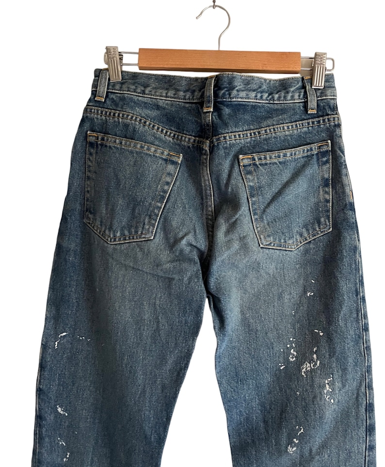 Puede incluir: Un par de pantalones vaqueros azules de mezclilla con salpicaduras de pintura blanca en las piernas. Los vaqueros tienen un estilo cl&aacute;sico de cinco bolsillos y est&aacute;n colgados de una percha de madera.