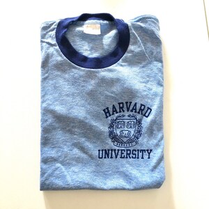 May include: A blue and navy blue t-shirt with a vintage look. The shirt has a ribbed crew neck and features a navy blue Harvard University logo with the text "HARVARD UNIVERSITY".