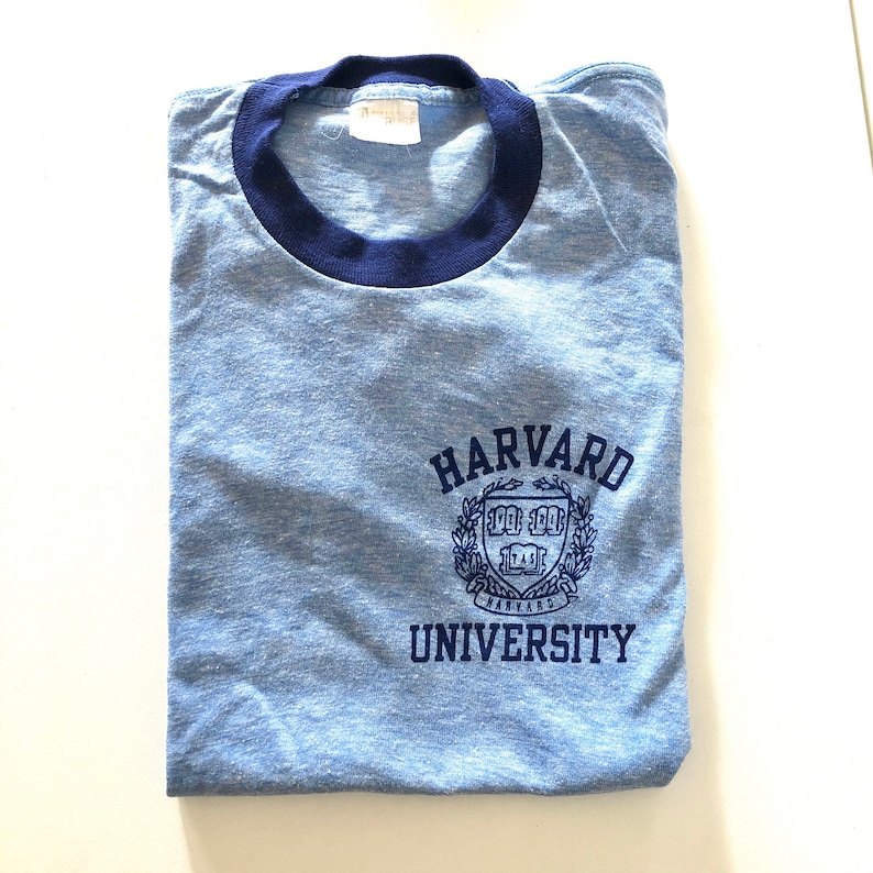 May include: A blue and navy blue t-shirt with a vintage look. The shirt has a ribbed crew neck and features a navy blue Harvard University logo with the text "HARVARD UNIVERSITY".