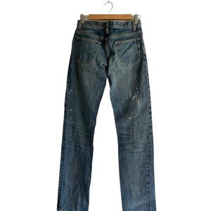 Puede incluir: Un par de pantalones vaqueros azules con salpicaduras de pintura blanca colgados de una percha de madera sobre un fondo blanco.