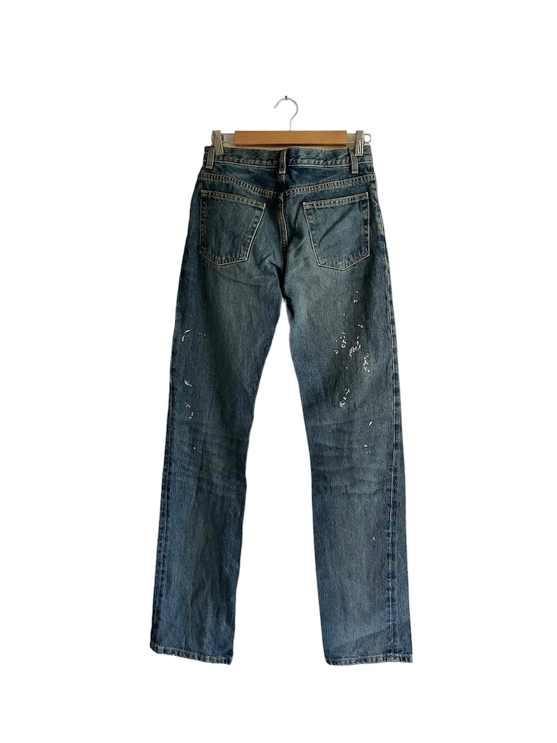 Puede incluir: Un par de pantalones vaqueros azules con salpicaduras de pintura blanca colgados de una percha de madera sobre un fondo blanco.