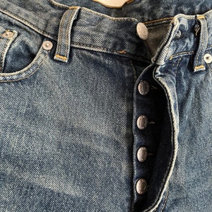 Puede incluir: Primer plano del cierre delantero de un par de pantalones vaqueros azules con cierre de bot&oacute;n. Los botones son plateados y tienen un dise&ntilde;o en relieve.