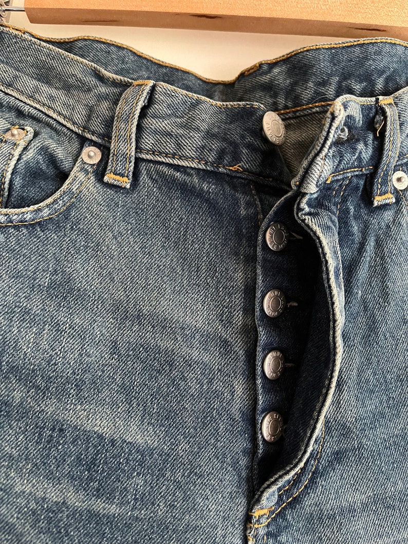 Puede incluir: Primer plano del cierre delantero de un par de pantalones vaqueros azules con cierre de bot&oacute;n. Los botones son plateados y tienen un dise&ntilde;o en relieve.