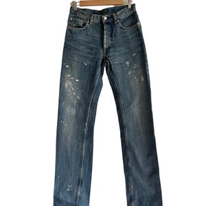 Puede incluir: Un par de pantalones vaqueros azules con salpicaduras de pintura blanca colgados de una percha de madera.