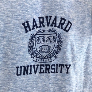 May include: A blue heather t-shirt with a black embroidered logo that says "Harvard University". The logo features a shield with a crest and a wreath of leaves.