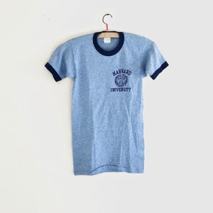 May include: A light blue t-shirt with a navy blue ringer and a navy blue embroidered Harvard University logo on the front.
