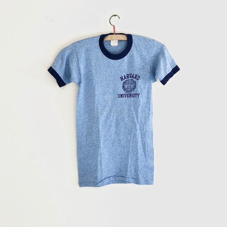 May include: A light blue t-shirt with a navy blue ringer and a navy blue embroidered Harvard University logo on the front.