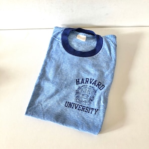 May include: A light blue t-shirt with a navy blue ringer neck. The shirt features a navy blue embroidered Harvard University logo on the front.