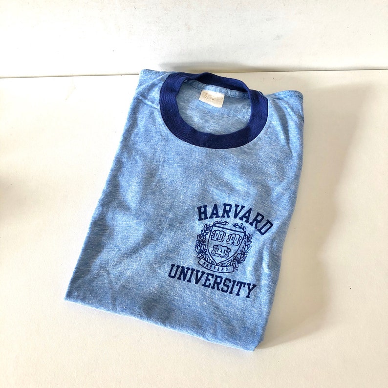 May include: A light blue t-shirt with a navy blue ringer neck. The shirt features a navy blue embroidered Harvard University logo on the front.