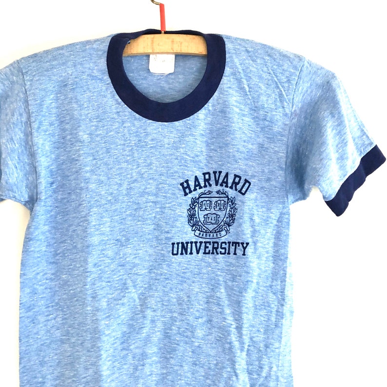 May include: A light blue t-shirt with a navy blue ringer. The shirt has a vintage look and features a navy blue embroidered Harvard University logo on the front.