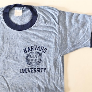 May include: A light blue t-shirt with a navy blue ringer. The shirt has a navy blue embroidered logo that says "HARVARD UNIVERSITY" with a crest in the middle.