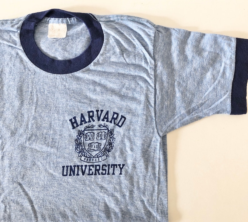 May include: A light blue t-shirt with a navy blue ringer. The shirt has a navy blue embroidered logo that says "HARVARD UNIVERSITY" with a crest in the middle.