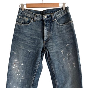 Puede incluir: Un par de pantalones vaqueros azules de mezclilla con salpicaduras de pintura blanca. Los vaqueros tienen un estilo cl&aacute;sico de cinco bolsillos y una pierna recta.