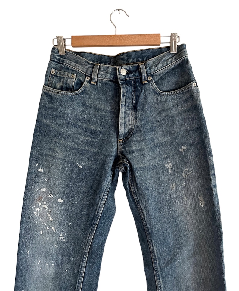 Puede incluir: Un par de pantalones vaqueros azules de mezclilla con salpicaduras de pintura blanca. Los vaqueros tienen un estilo cl&aacute;sico de cinco bolsillos y una pierna recta.