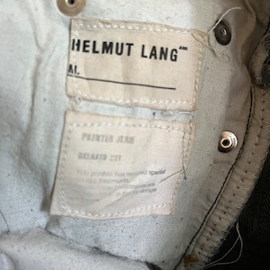 Puede incluir: Una etiqueta blanca con el texto "HELMUT LANG" y "PAINTER JEANS RELAXED FIT" en un par de jeans azul oscuro.