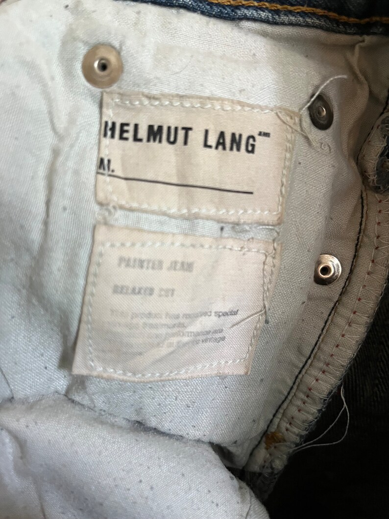 Puede incluir: Una etiqueta blanca con el texto "HELMUT LANG" y "PAINTER JEANS RELAXED FIT" en un par de jeans azul oscuro.