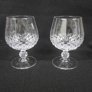 Peut inclure: Deux verres à cognac en cristal transparent. Les bols présentent un motif taillé en diamant et les pieds sont ornés d'une boule décorative. Les verres reposent sur une base ronde. Le fond est sombre.