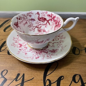 Peut inclure: Une tasse et une soucoupe blanches avec un motif floral et d'oiseaux rouge. La tasse a une anse incurvée et une bordure dorée. La soucoupe a un motif assorti et une bordure dorée. L'ensemble est sur une surface en bois.