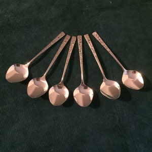 May include: Six silver teaspoons with ornate designs on the handles. The spoons are arranged in a circle on a green surface.