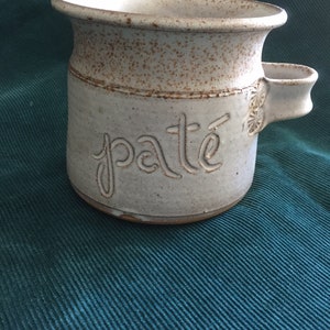 May include: A white ceramic crock with a brown glaze and the word "pate" written on the front. The crock has a small handle on the side.
