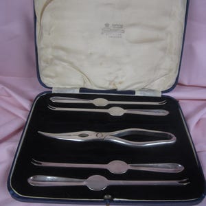 May include: A blue velvet box with a white satin lining contains a set of four silver oyster forks and a pair of silver oyster crackers. The box has a label that reads "Sheffield England, Thomas Bradbury & Sons, Makers, Sheffield."