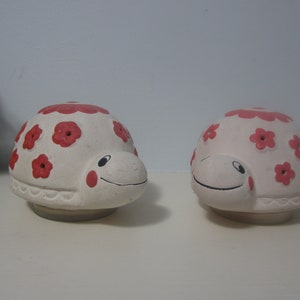 May include: Two white ceramic turtle figurines with red flower designs. The turtles have a smiling face and are facing each other.