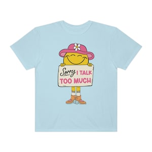 以下が含まれることがあります： 薄い青色のTシャツに、ピンクの帽子をかぶって「ごめんなさい、話しすぎます」と書かれた看板を持っている、漫画の笑顔の顔のデザインが描かれています。