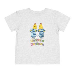 May include: A gray t-shirt with a graphic of two yellow bananas wearing blue and white striped pajamas. The text "Bananas in Pyjamas" is printed in colorful block letters below the bananas.