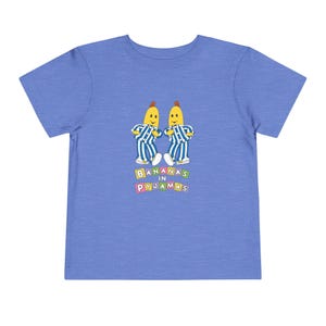 May include: A light blue toddler t-shirt featuring a graphic of two yellow bananas in blue and white striped pajamas. The text "BANANAS IN PYJAMAS" is printed in colorful block letters below the bananas.