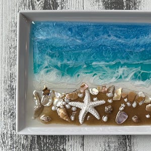 May include: A rectangular serving tray with a beach scene. The tray features a blue and white ocean design, a sandy shore, and seashells. A white starfish is on the shore. The tray is white with a glossy finish.
