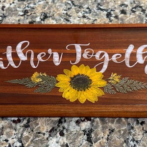 May include: A rectangular wooden serving tray with the words "gather together" in white script. A yellow sunflower and dried greenery are centered below the text. The tray is a rich brown color.
