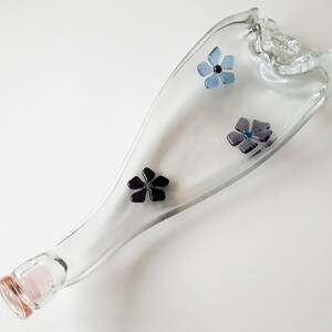 May include: A clear glass bottle with a pink stopper and a unique, curved shape. The bottle is decorated with four small, colourful flowers.