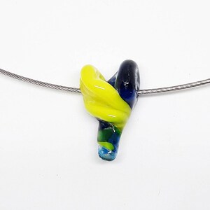 May include: A yellow, blue, and green glass pendant hangs from a silver chain. The pendant is shaped like a swirl or a knot.