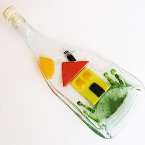 May include: A glass bottle with a house design made of colourful glass pieces. The house is yellow with red and orange accents. The bottle is filled with green glass pieces.