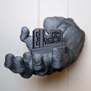 May include: A dark gray, geometric-patterned object with a honeycomb design is held in a textured, gray hand. The object has a rectangular base and a series of vertical and horizontal lines. The hand is the focal point, with detailed texture.