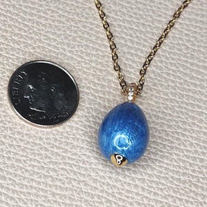 May include: A gold-colored chain necklace with a blue pendant shaped like an egg. The pendant has a metallic finish and features a gold-colored number 8. A dime is next to the necklace.