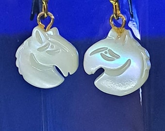 May include: A pair of earrings with a horse head design. The earrings are white with iridescent highlights and gold-colored hooks. The horse heads have carved details, including eyes and a mouth. The background is a solid, dark blue.