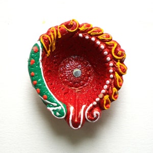 May include: A red clay diya with green, white, and yellow painted designs. The diya is a traditional Indian oil lamp used during Diwali.