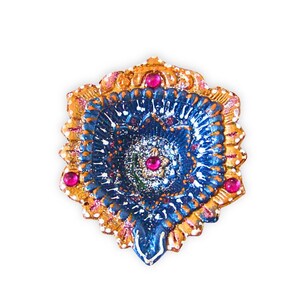 May include: A decorative diya oil lamp featuring a scalloped edge painted in metallic gold with white accents. The center is a deep blue with intricate floral patterns, glittery details, and three embedded bright pink circular gemstones.