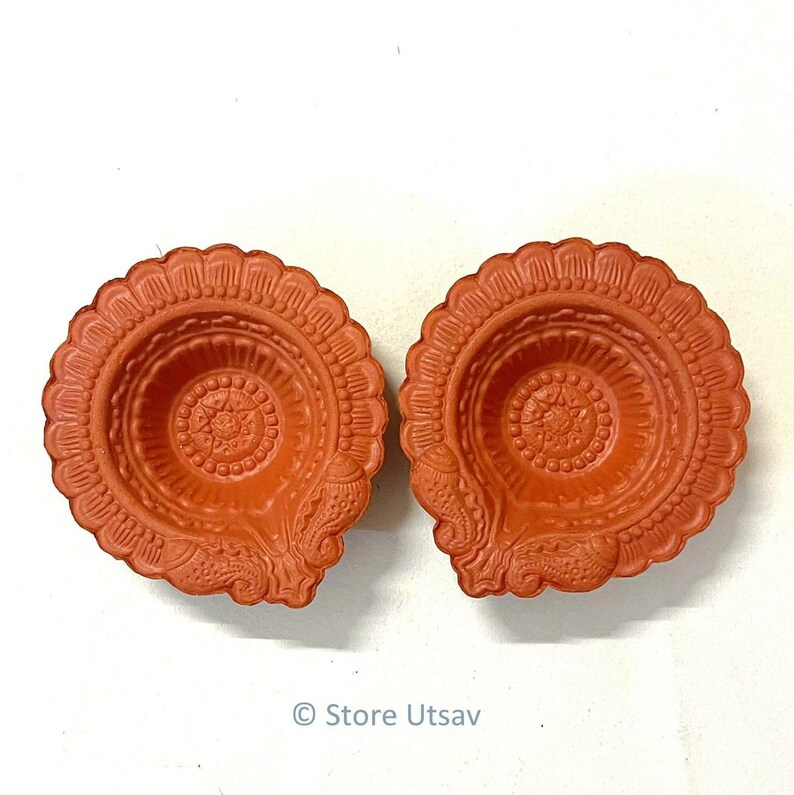 Pode incluir: Duas l&acirc;mpadas de &oacute;leo decorativas de cor terracota. Cada l&acirc;mpada apresenta uma borda ondulada e padr&otilde;es em relevo intrincados, incluindo um design floral central e motivos laterais detalhados. As l&acirc;mpadas s&atilde;o projetadas para conter &oacute;leo e um pavio.