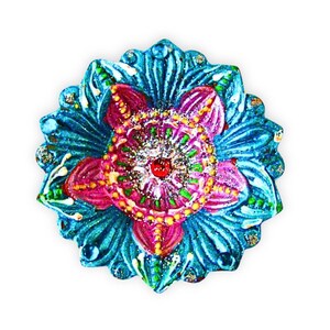 May include: A circular decorative diya lamp featuring a vibrant floral pattern with layered petals in shades of turquoise, magenta, and bright green. The center is accented with a small red bead and silver glitter, with additional clear crystal embellishments.
