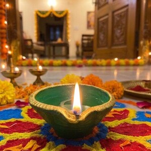 May include: A green clay oil lamp with a gold-painted rim and a lit flame, resting on a colorful circular floor pattern made of colored powder. The scene includes scattered orange and yellow marigold flowers and small brass bowls nearby.